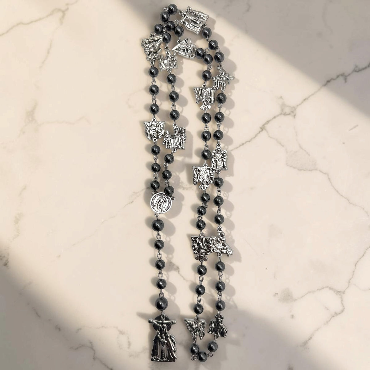 Vertical view of Way of the Cross chaplet with dark gray hematite beads, silver station medals, Holy Face centerpiece, and crucifix