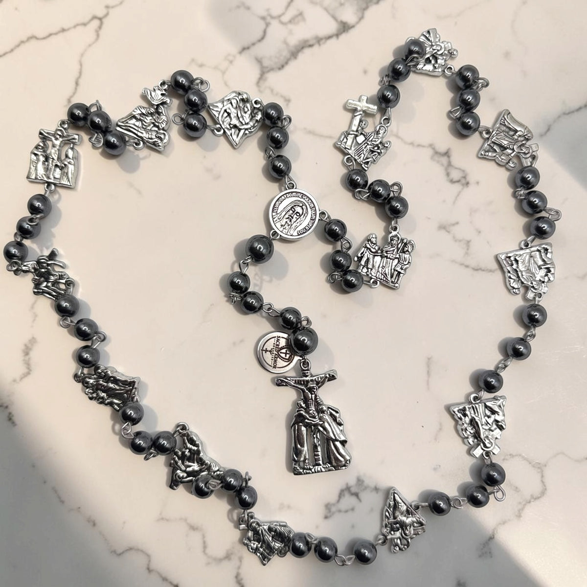Way of the Cross chaplet arranged in a heart shape with dark gray hematite beads, silver station medals, Holy Face of Jesus centerpiece, and silver crucifix