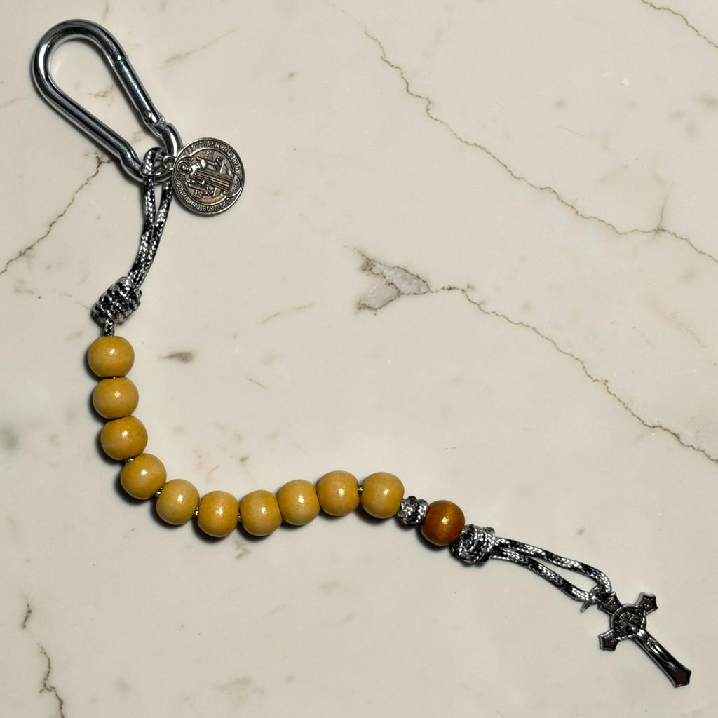 Saint Benedict decade rosary laid out with beige wood beads, tan Our Father bead, black and white paracord, carabiner, medal, and silver-tone crucifix