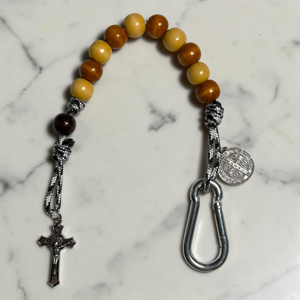 Close-up of Saint Benedict decade rosary with alternating beige and warm brown wood beads, dark brown Our Father bead, black and white paracord, medal, carabiner, and crucifix