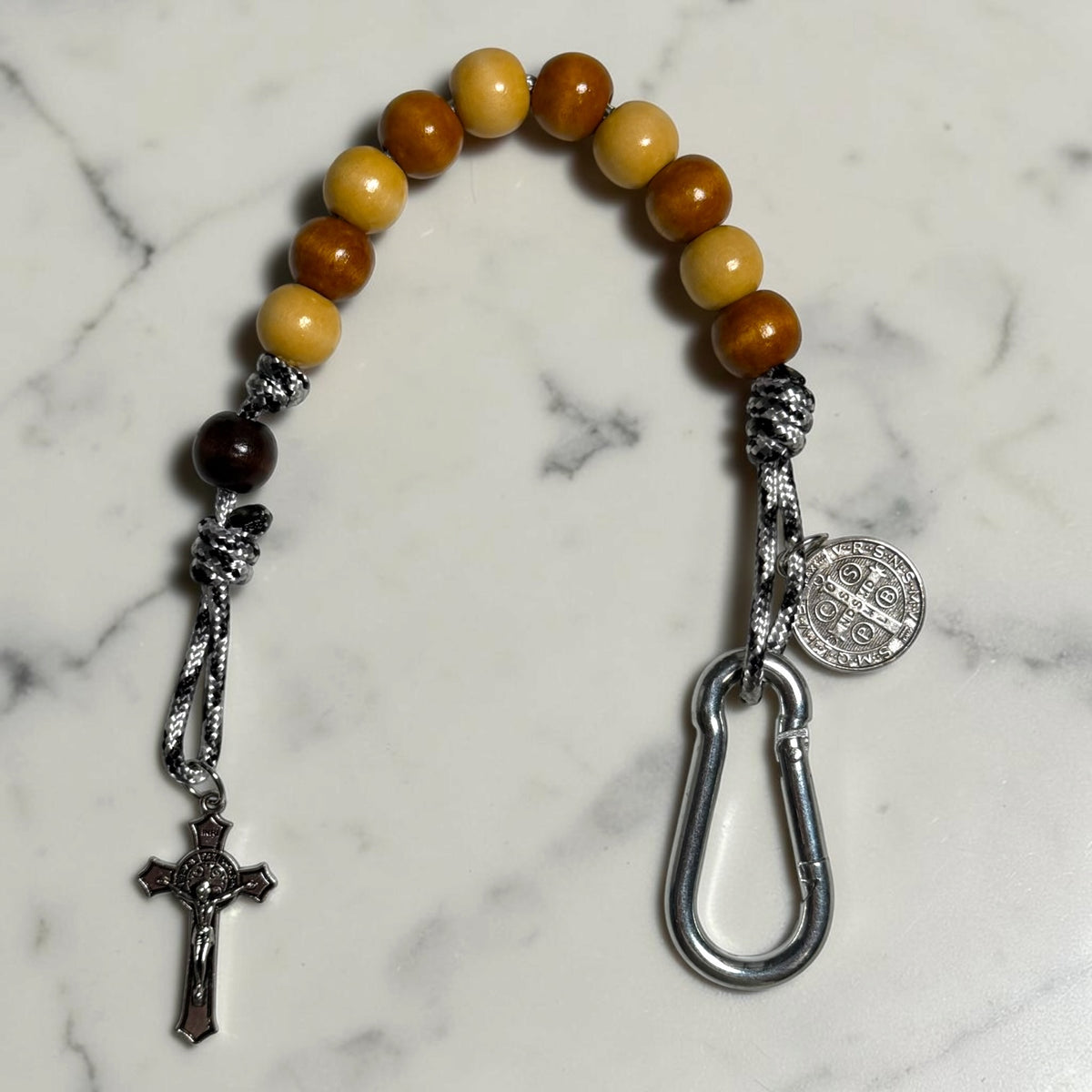 Close-up of Saint Benedict decade rosary with alternating beige and warm brown wood beads, dark brown Our Father bead, black and white paracord, medal, carabiner, and crucifix