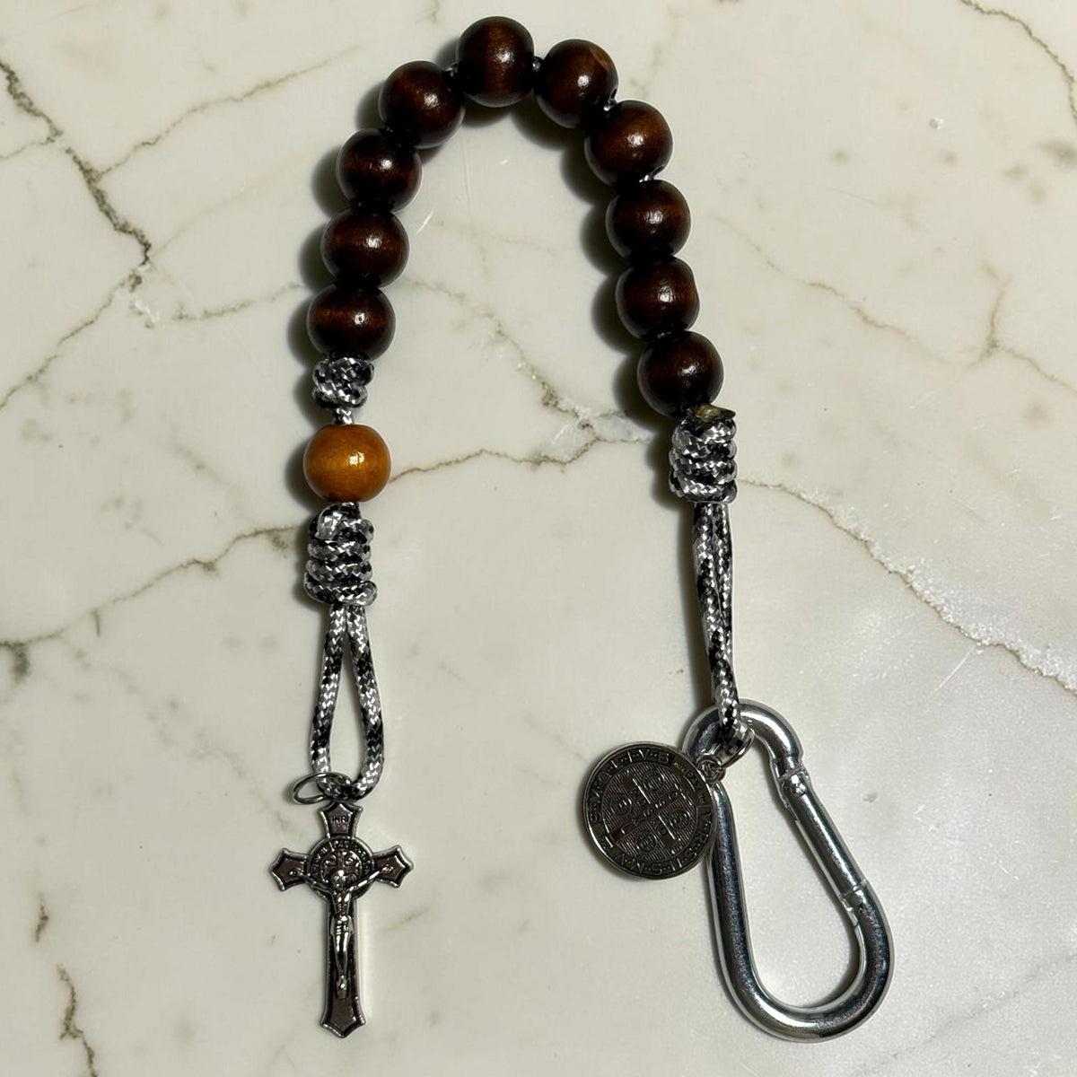 Saint Benedict decade rosary with dark brown wood beads, tan Our Father bead, black and white paracord, silver-tone carabiner, medal, and crucifix