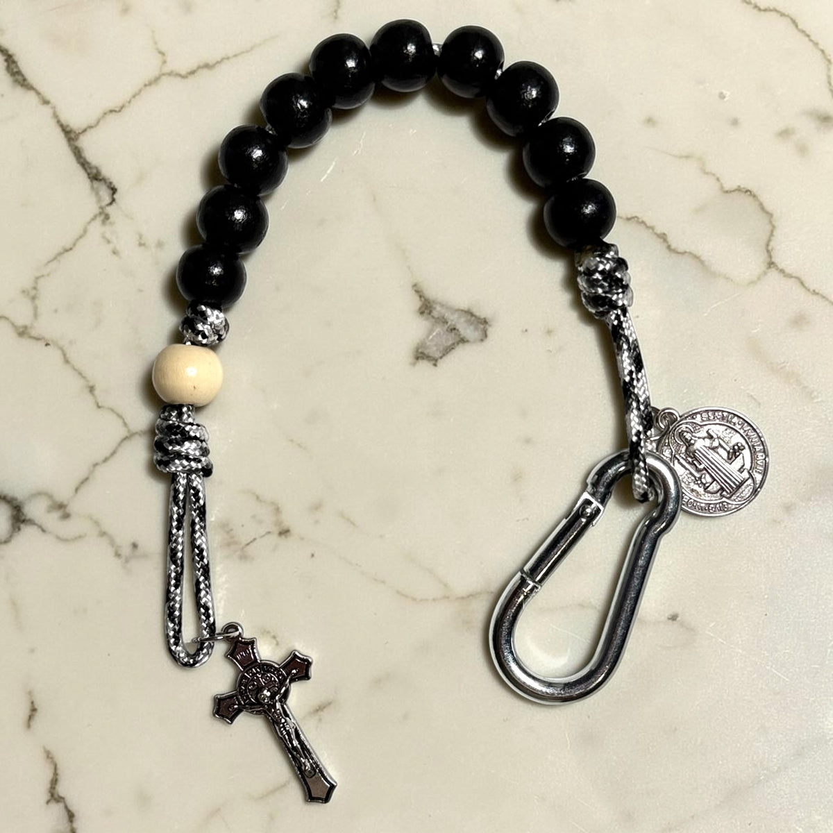 Close-up Saint Benedict decade rosary with black wood beads, white Our Father bead, black and white paracord, carabiner, medal, and crucifix