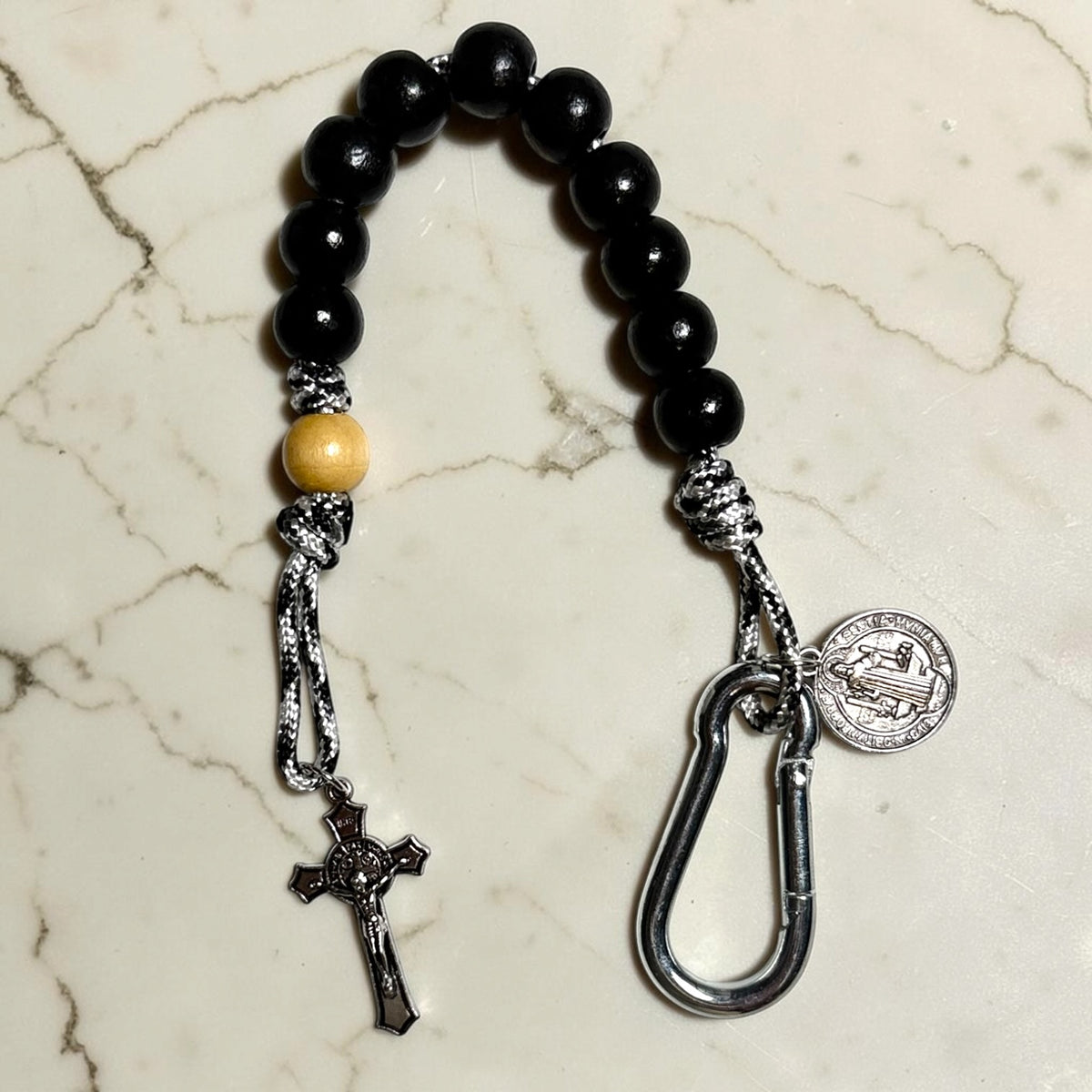 Saint Benedict decade rosary with black wood beads, beige Our Father bead, black and white paracord, silver-tone carabiner, medal, and crucifix