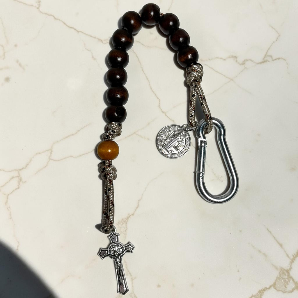 Close-up of Saint Benedict decade rosary with dark wood beads, desert camo paracord, silver medal, carabiner, and crucifix
