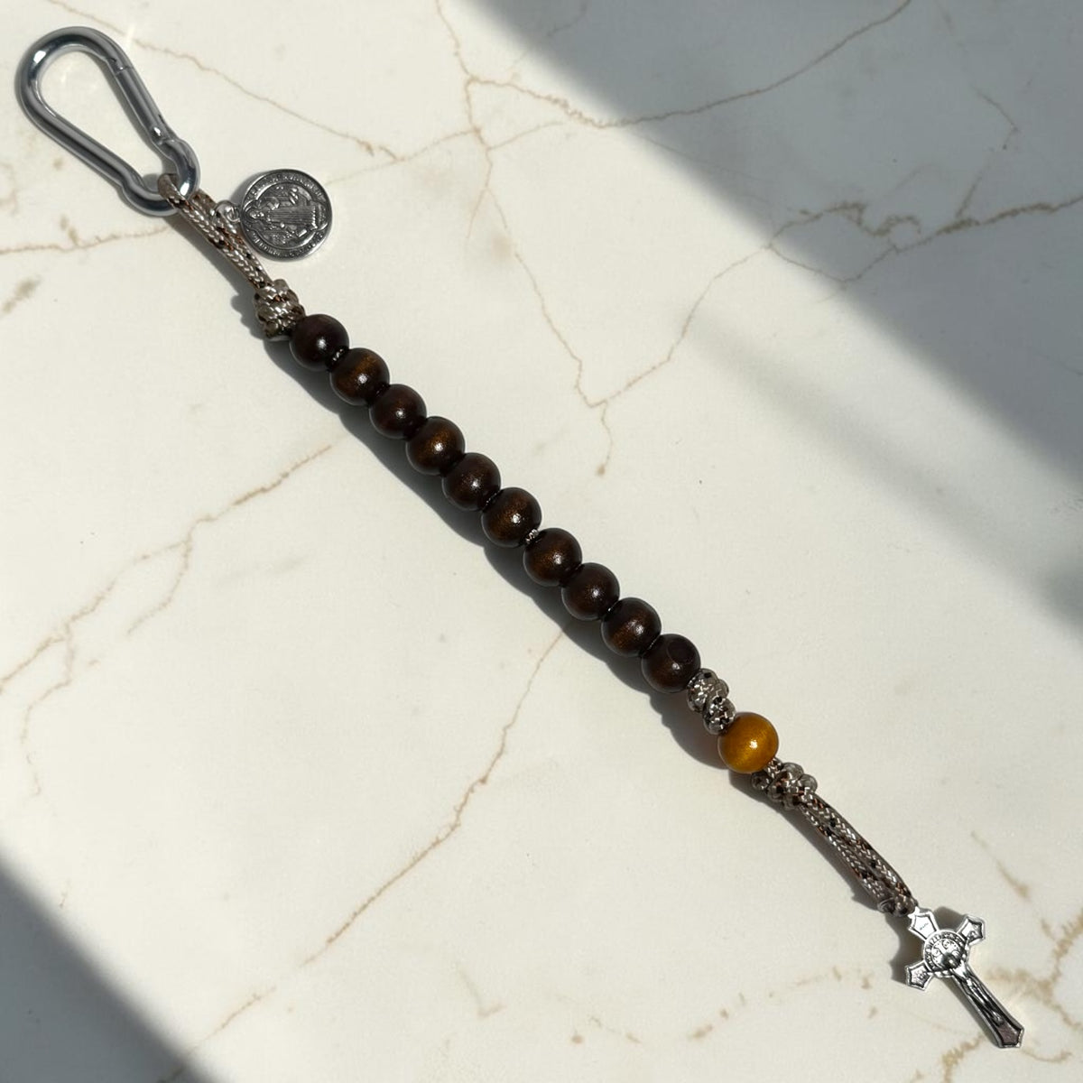 Saint Benedict decade rosary laid out with dark wood beads, warm accent bead, desert camo paracord, and silver crucifix