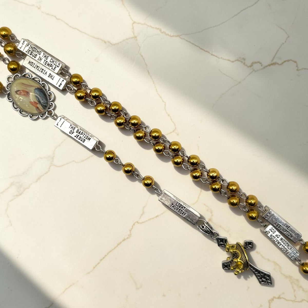 Close-up of silver and gold guided mystery rosary showing mystery plaques, gold beads, Marian centerpiece, and crown crucifix
