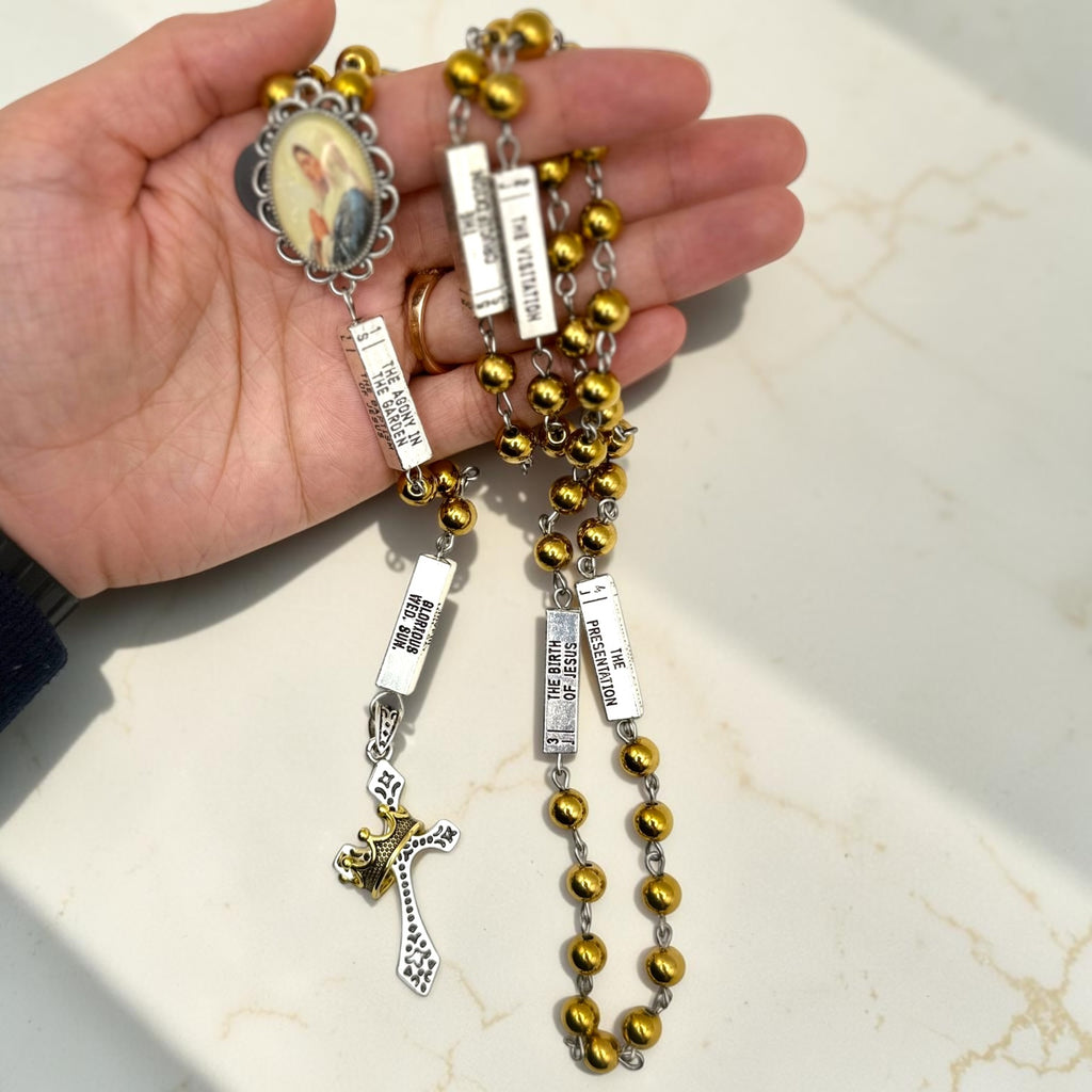 Handheld silver and gold guided mystery rosary showing mystery plaques, gold beads, Marian centerpiece, and crown crucifix