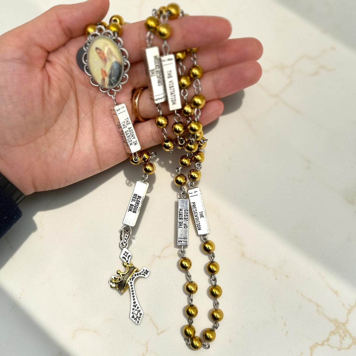 Handheld silver and gold guided mystery rosary showing mystery plaques, gold beads, Marian centerpiece, and crown crucifix