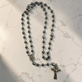 Dark gray hematite Miraculous Medal rosary arranged on marble with silver-tone chain, larger silver Our Father beads, Marian centerpiece, and ornate crucifix