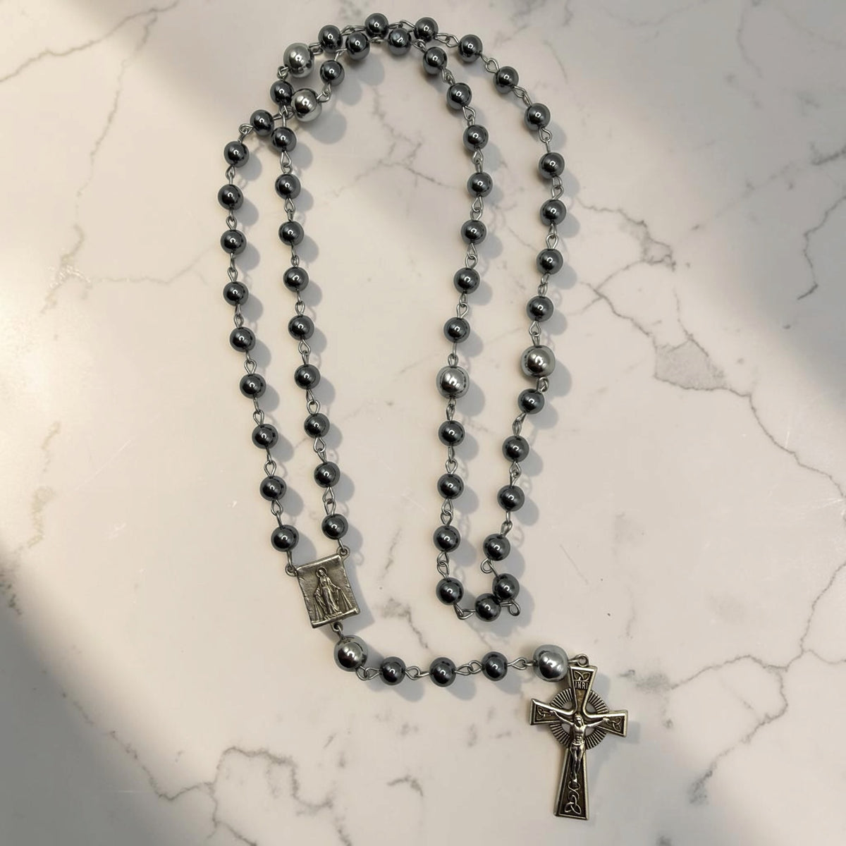Dark gray hematite Miraculous Medal rosary arranged on marble with silver-tone chain, larger silver Our Father beads, Marian centerpiece, and ornate crucifix