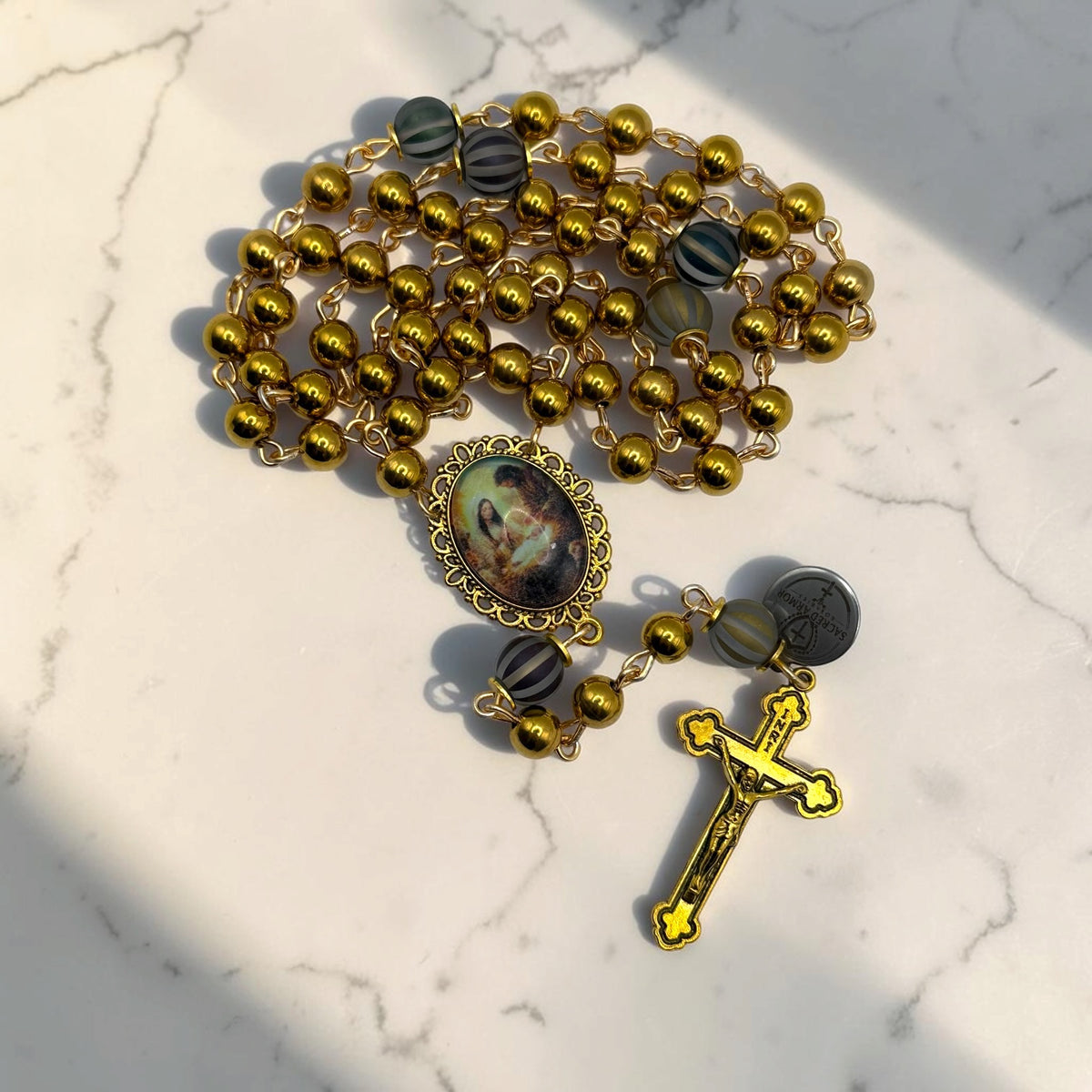 Close-up of gold Holy Family rosary with striped accent beads, ornate Holy Family centerpiece, Sacred Armor medal, and gold crucifix