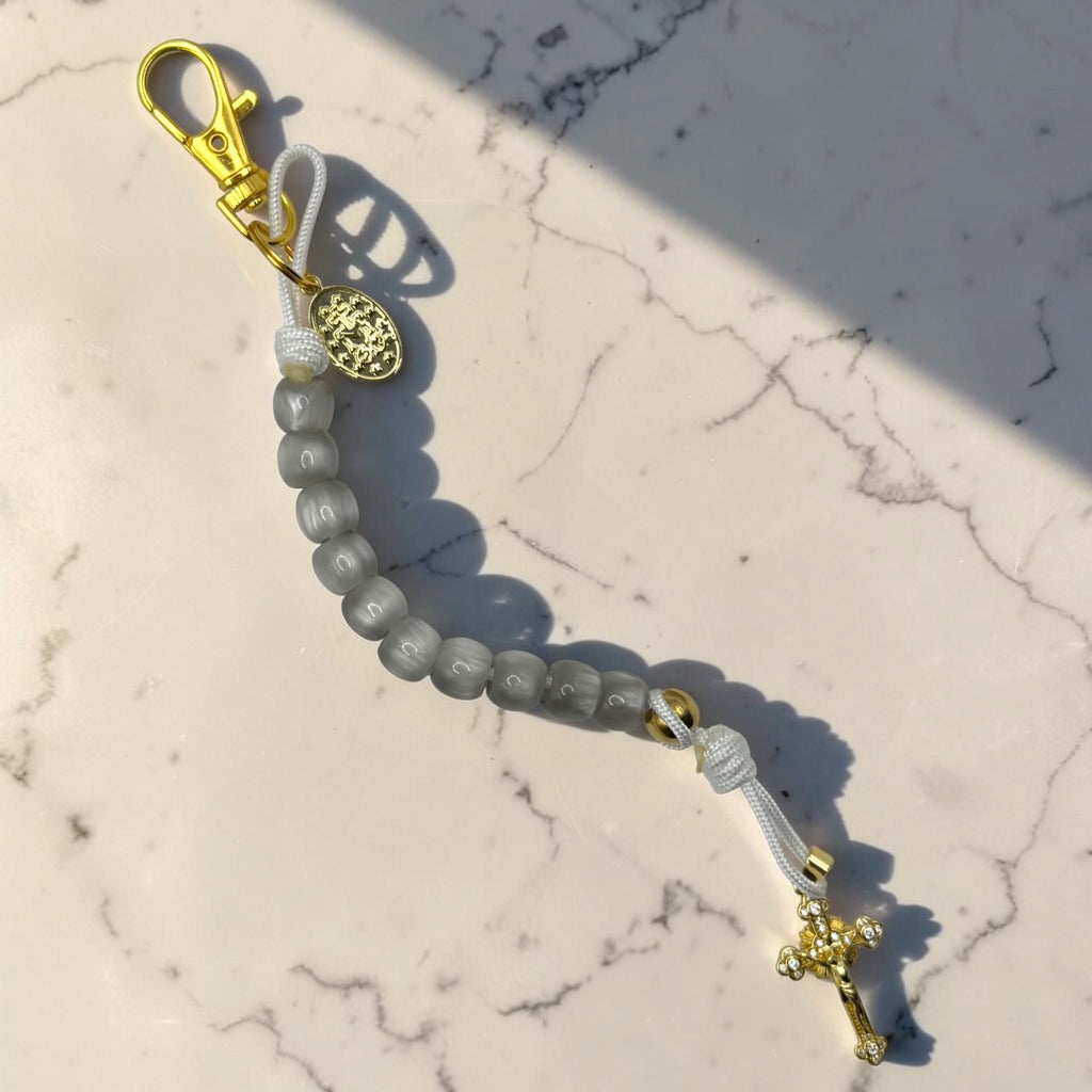 Gray decade rosary in sunlight with white cord, gold-tone Marian medal, lobster clasp, and crystal crucifix