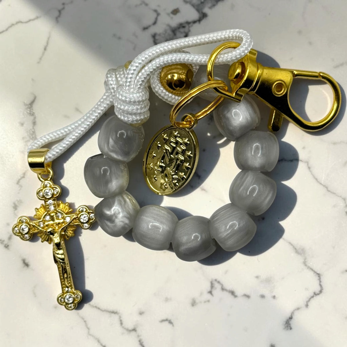Close-up gray decade rosary with glossy gray beads, white cord, gold-tone Marian medal, lobster clasp, and crystal crucifix
