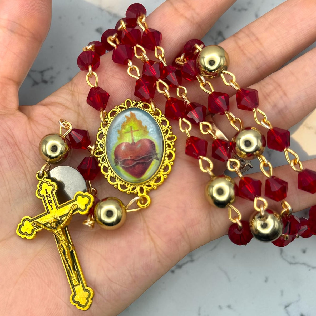 Hand holding red faceted crystal Catholic rosary with gold-tone beads and Sacred Heart centerpiece