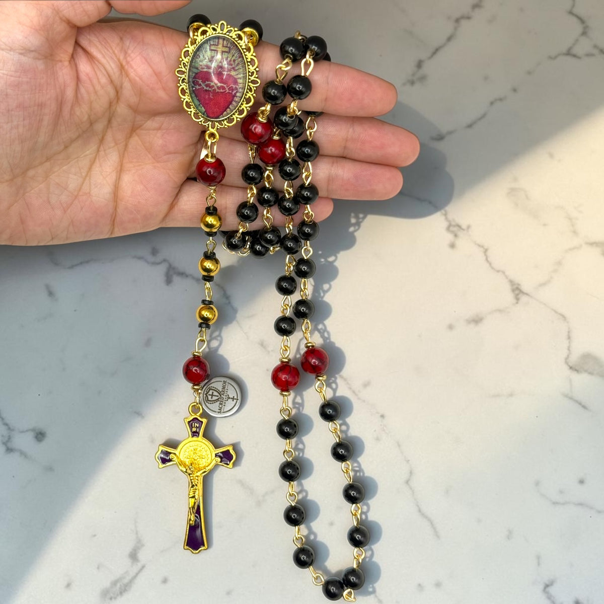 Hand holding black and red Sacred Heart rosary with gold-tone chain, gold accent beads, and purple-accented crucifix