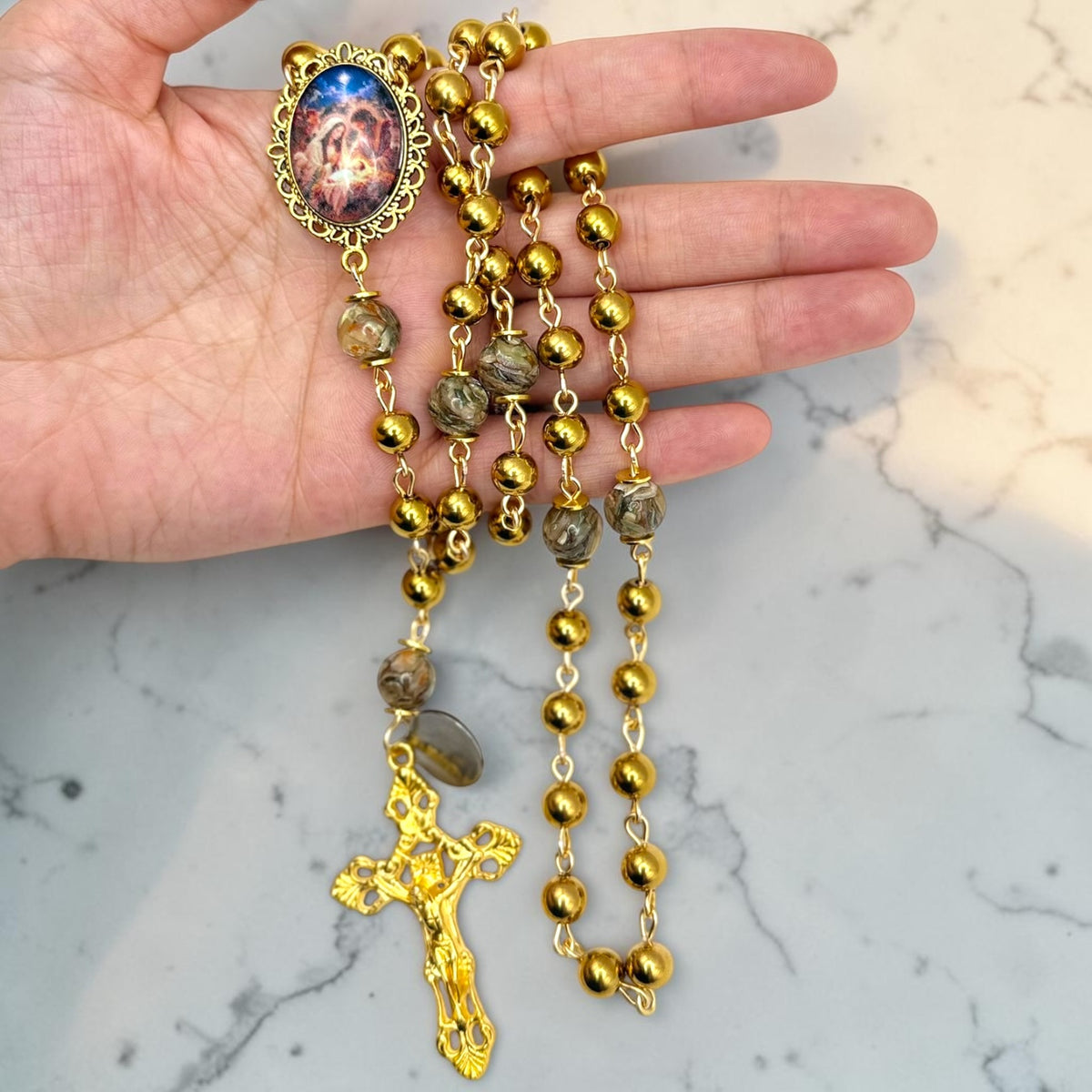 Hand holding gold-tone Catholic rosary with marbled glass accent beads, devotional centerpiece, and ornate gold crucifix